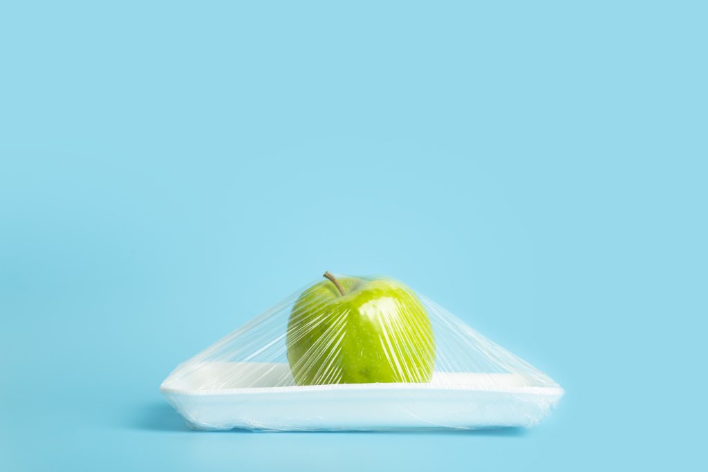 Fruits in plastic packaging from the supermarket are minimal. Apple in cellophane and non-degradable plastic on a blue background. Biodegradable product packaging, environmental protection, nature pollution. Fruits in plastic packaging from the supermarket are minimal. Apple in cellophane and non-degradable plastic on a blue background. Biodegradable product packaging, environmental protection, nature pollution. High quality photo